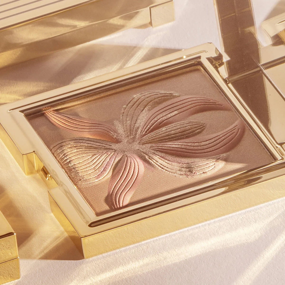 Gold-toned rectangular compact with bronze powder featuring a raised orchid design in varying shades of bronze and pinkish-br