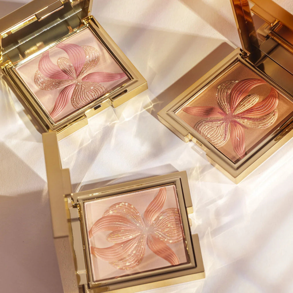 Square gold-toned compact with bronze pressed powder featuring a raised orchid design with pink and gold-toned glitter accent