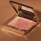 Open gold-toned compact with mirror, revealing light pink powder featuring a coral-pink orchid design with gold-toned glitter