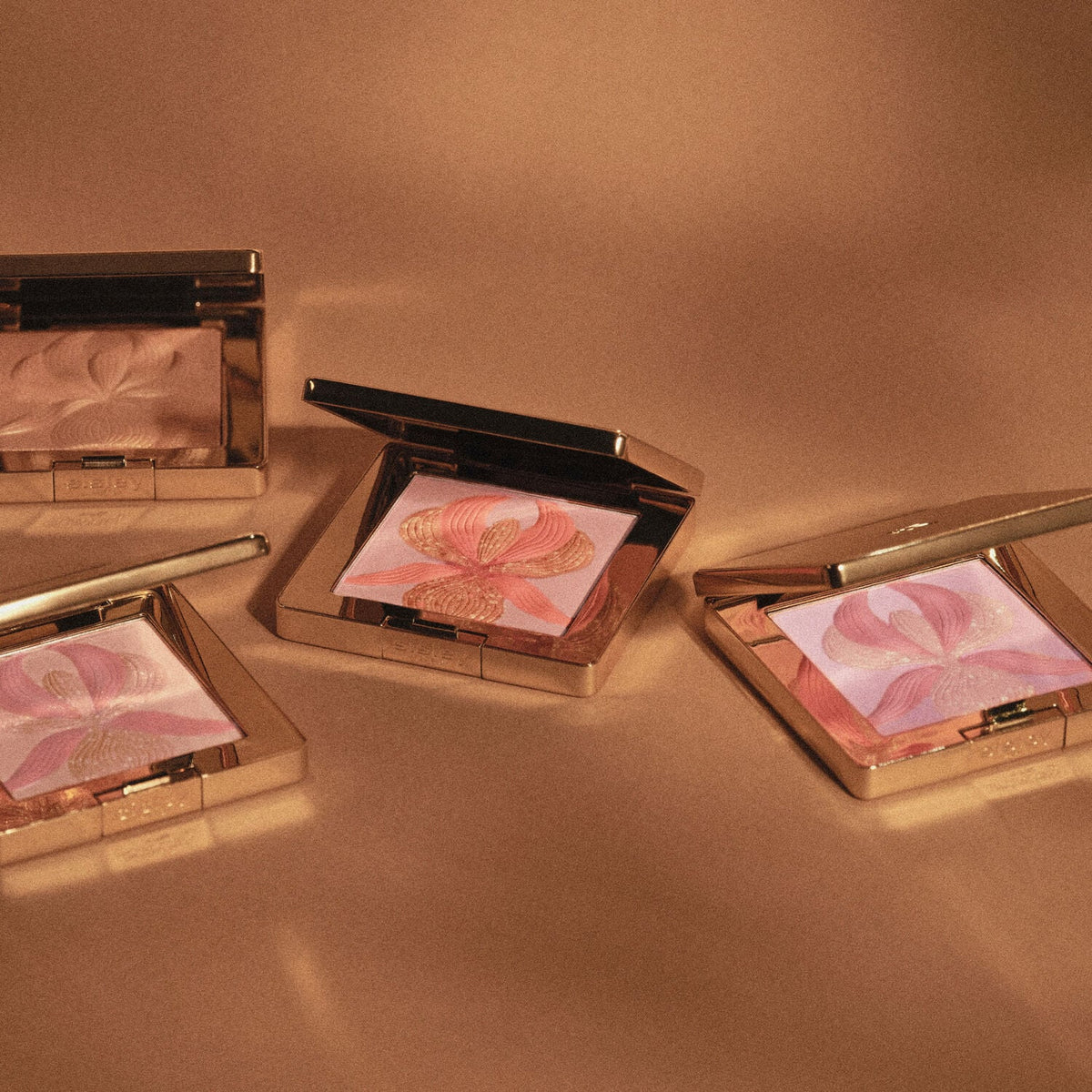 Gold-toned square compact with an open lid, revealing pressed powder with an embossed pink, peach, and gold-toned orchid desi