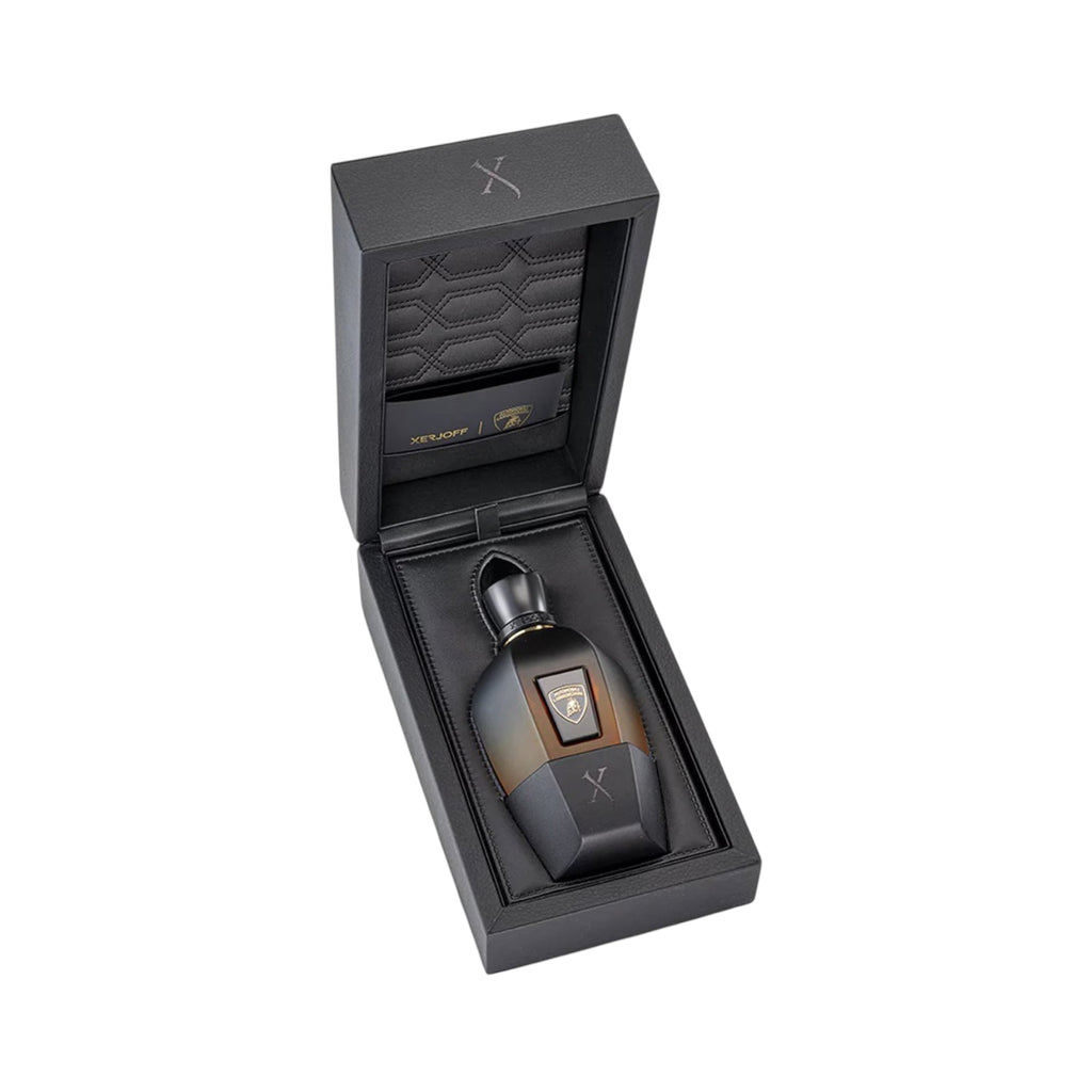 Black hinged box open to reveal a perfume bottle. The bottle is dark brown to amber ombre with a black cap, gold-toned emblem