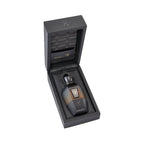 Black hinged box open to reveal a perfume bottle. The bottle is dark brown to amber ombre with a black cap, gold-toned emblem