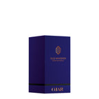 A luxury bottle of OJAR Oud Whisper Extrait de Parfum. The cylindrical bottle is crafted from deep, translucent midnight-blue glass and features the OJAR logo and fragrance name in a vibrant copper-orange foil. It is topped with a reflective copper-toned metallic cap, presented against a clean white background.