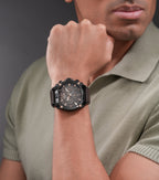 Black watch with a black multi-function dial, rose-gold-toned accents, and a textured brown leather strap.