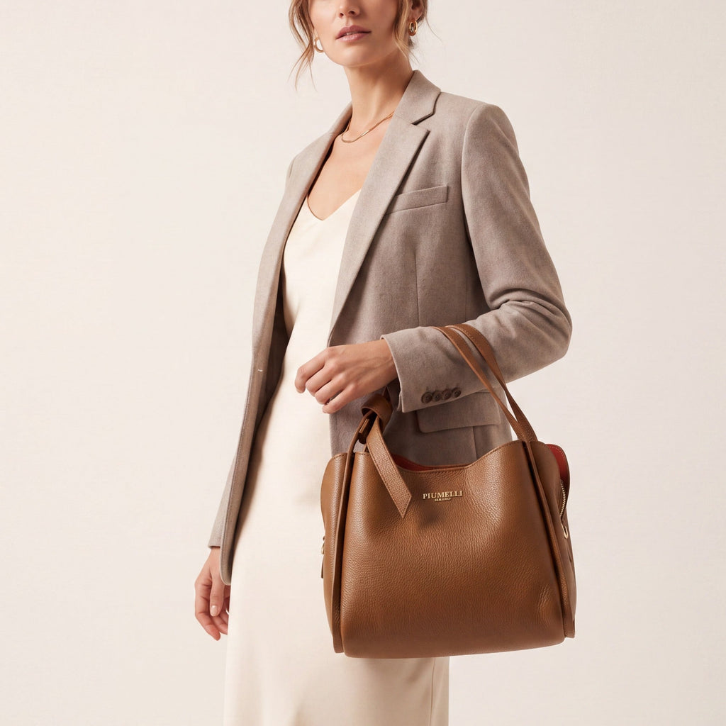 Warm brown pebbled leather tote bag with two top handles, gold-toned PIUMELLI logo, red interior, and decorative side strap.
