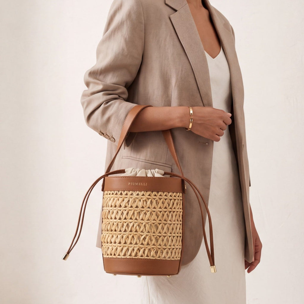 Brown leather and woven straw bucket bag with a cream drawstring closure, gold-toned hardware, and "PIUMELLI" logo.