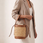Brown leather and woven straw bucket bag with a cream drawstring closure, gold-toned hardware, and "PIUMELLI" logo.