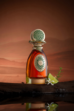 Amber perfume in clear bottle, gold-toned base. Gold-toned oval with green mosaic, central symbol. Gold-toned ornate neck, sp
