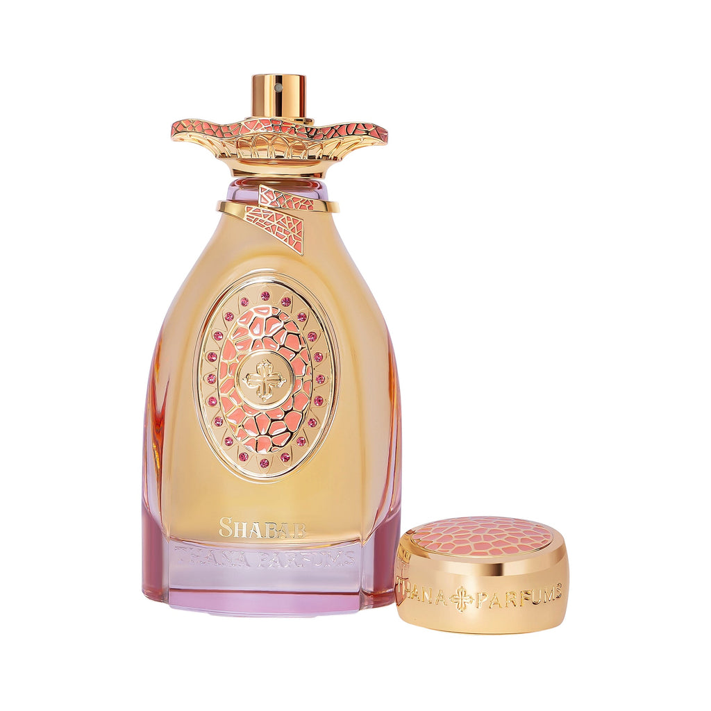 Gold-toned, curved glass perfume bottle with a pink-purple base, an oval coral-pink emblem with gems, and a matching ornate c