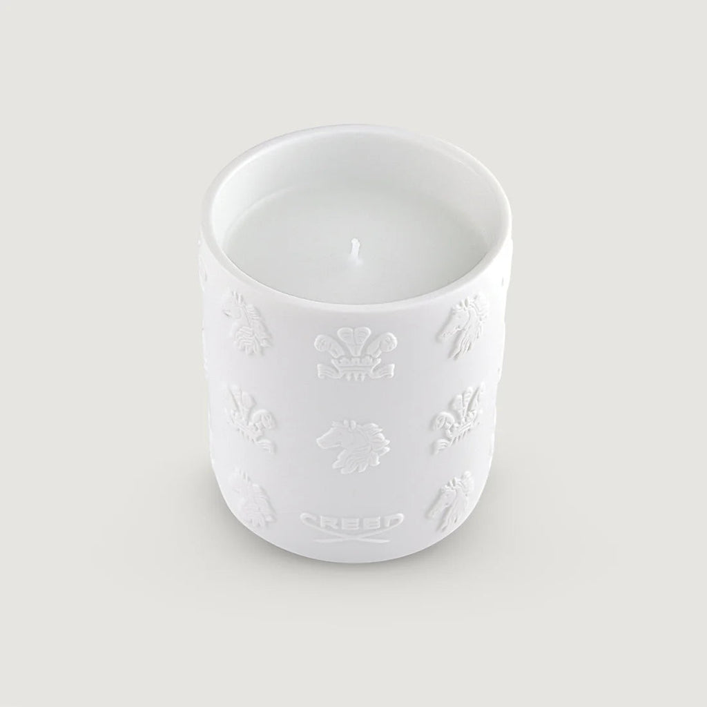 White candle in a round, white ceramic holder with embossed crests, horse heads, and the word 'CREED' on the side.