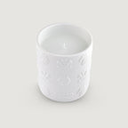 White candle in a round, white ceramic holder with embossed crests, horse heads, and the word 'CREED' on the side.