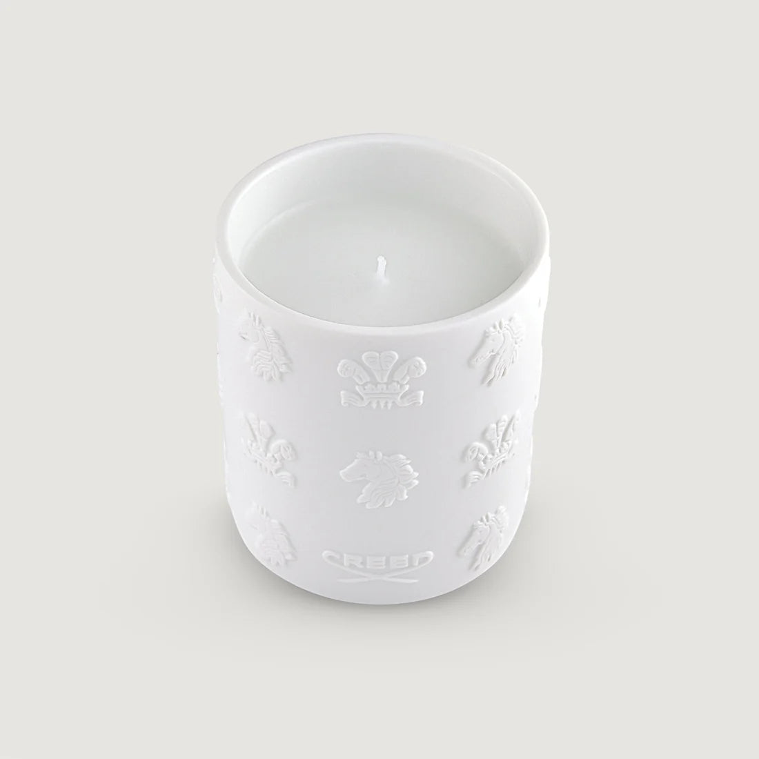 White ceramic candle holder with embossed crown, horse, and plume designs. White wax and a single wick are visible inside. Th