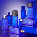 Rectangular clear glass bottle with blue-purple liquid, gold-toned spray nozzle. White text reads 'Hair Fragrance with Kerati